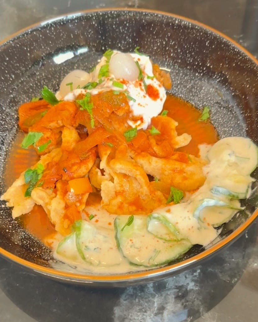 Chicken paprikash over nokedli in a dark bowl, with sour cream, parsley and creamy cucumber salad