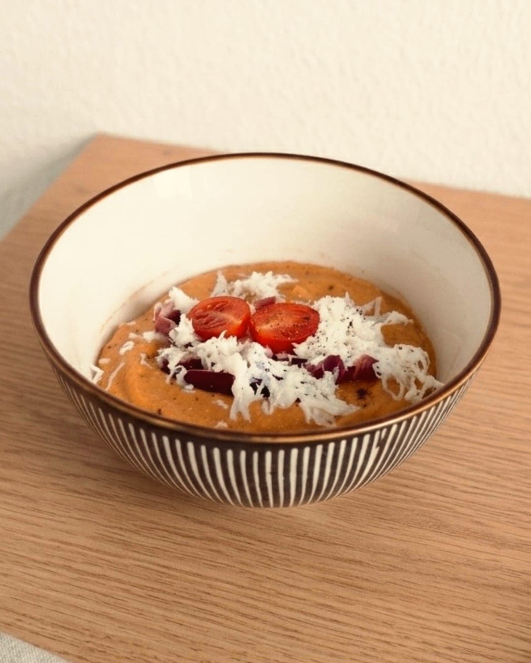 A striped bowl of coral salmorejo topped with grated egg, jamón and halved cherry tomatoes